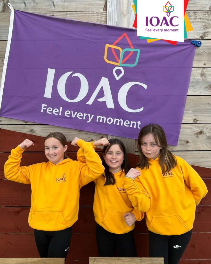 Three girls in yellow hoodies flexing by a purple 'IOAC' banner.