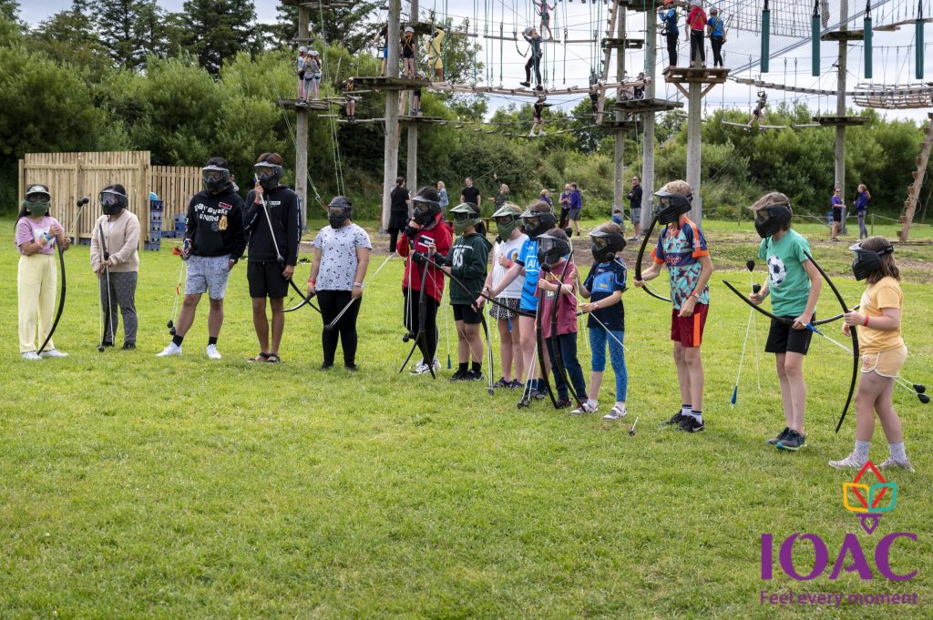 Children in line holding bows, wearing masks, outdoors with rope course background.