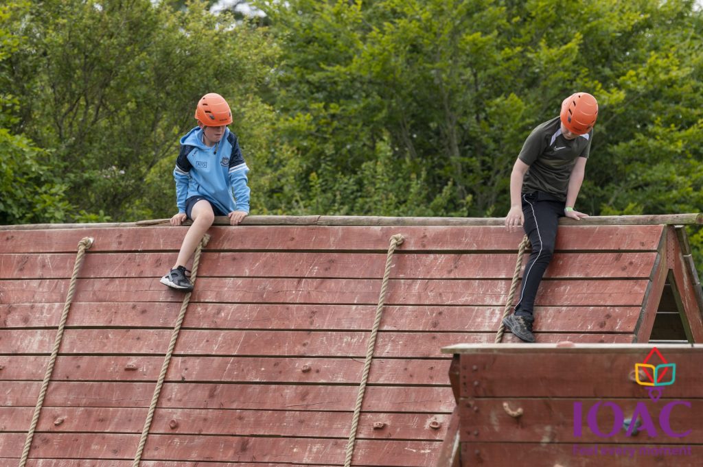Two people climb a wooden obstacle wall outdoors, wearing helmets.
