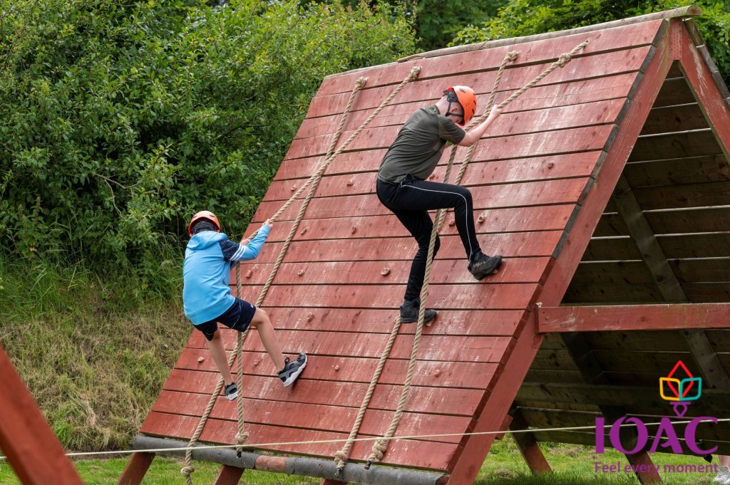 Two people climb a wooden obstacle using ropes outdoors.