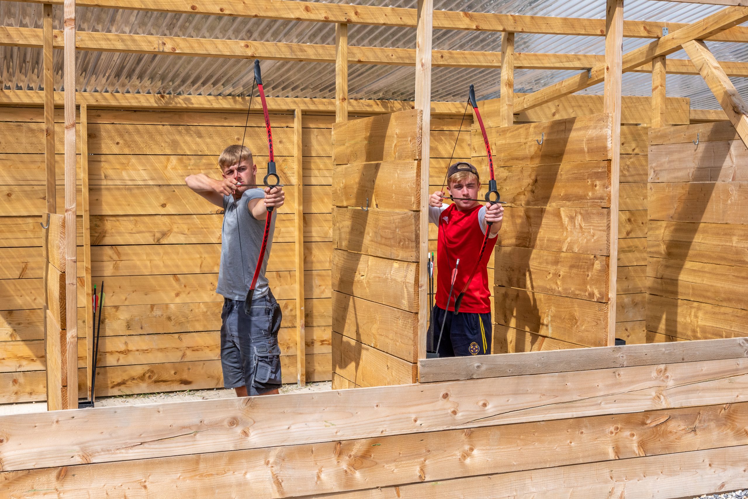 Two boys practicing archery in an outdoor wooden enclosure.
