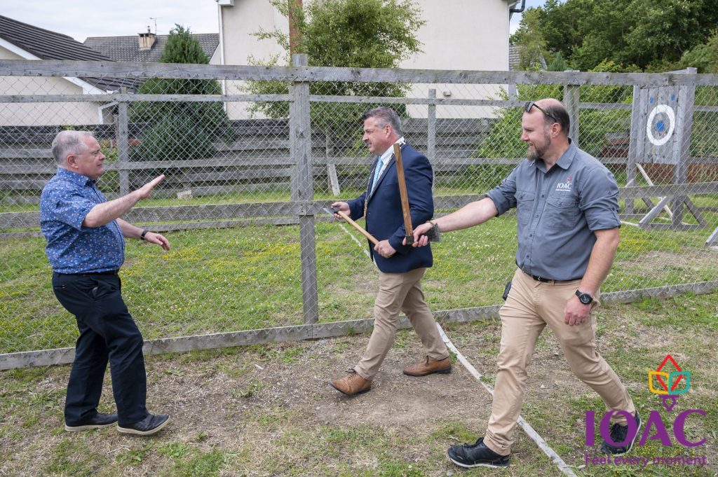 Three men standing outside near a wooden fence, two holding bows and arrows.