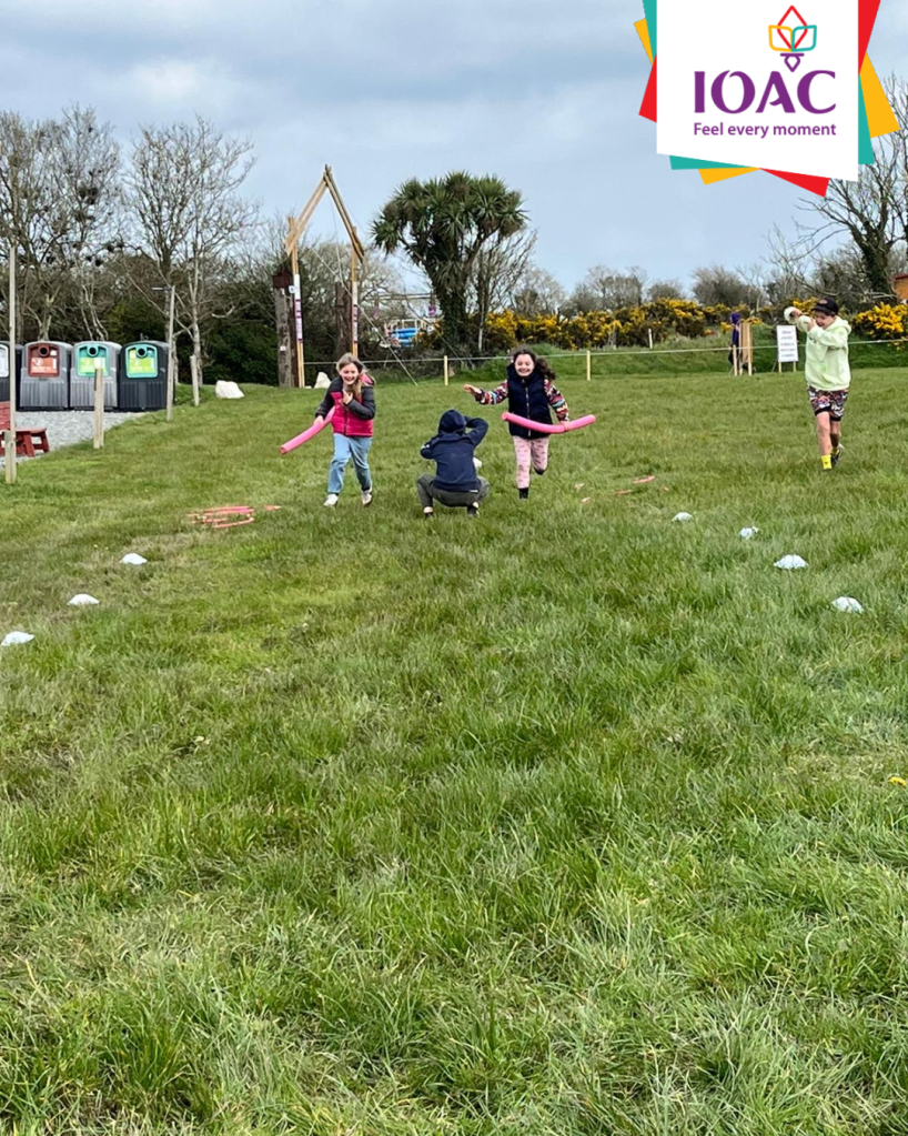 Children running on grass towards a standing adult, with trees and a logo in the background.