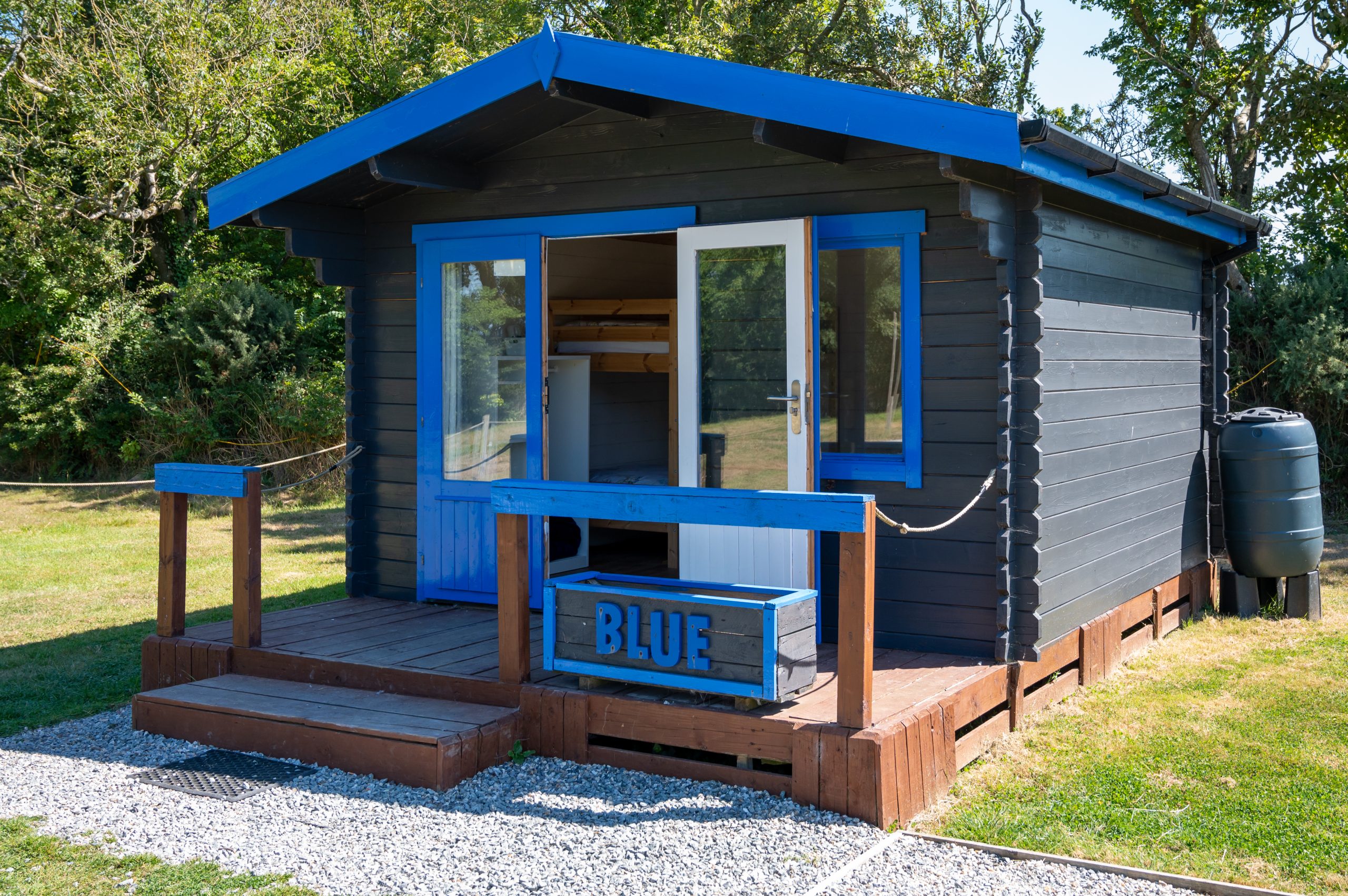 Small blue cabin with a porch in a grassy area surrounded by trees.