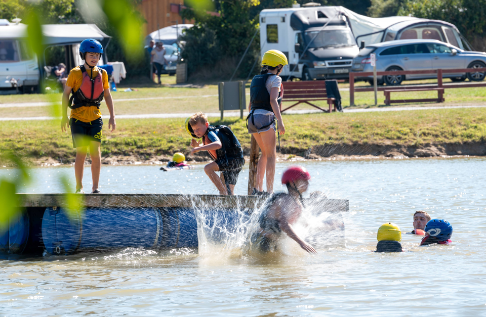 Children in helmets and life vests playing on a lake dock with splashing water.