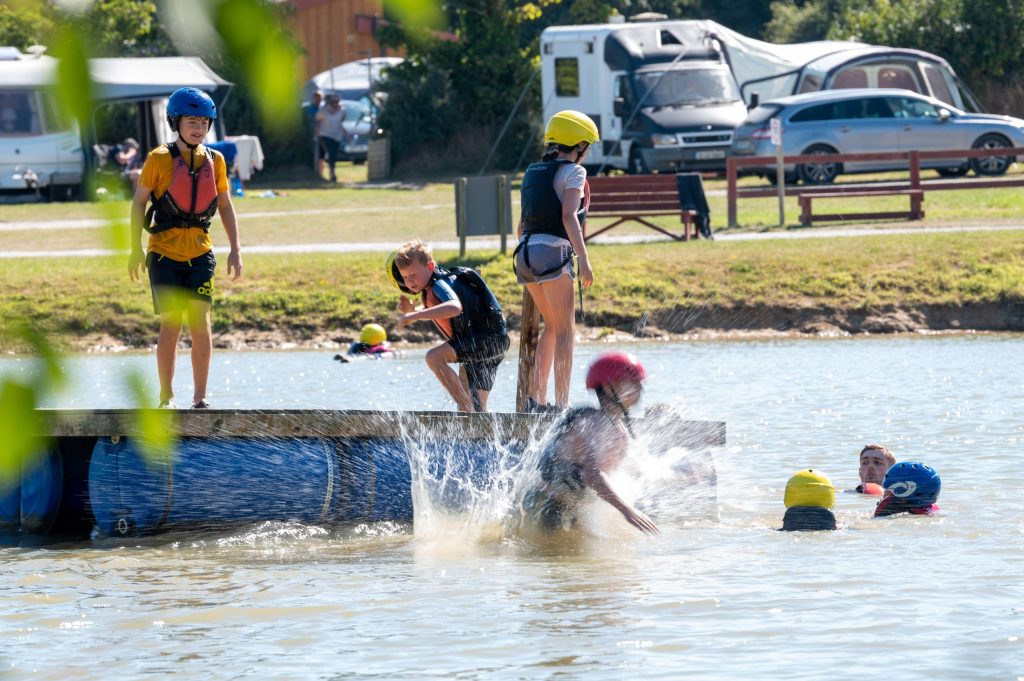 Children in helmets and life vests play on a lake dock, with some in water and a splash visible.