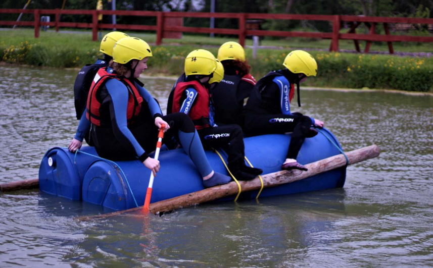 Four people in helmets on a homemade barrel raft in a pond.