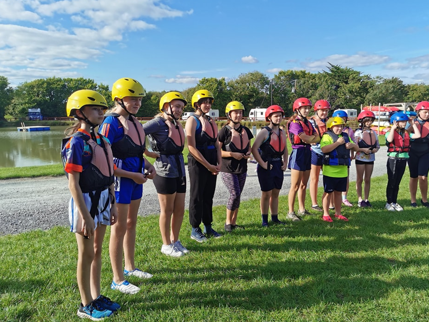 Group of people in helmets and life vests standing on grass by a lake.