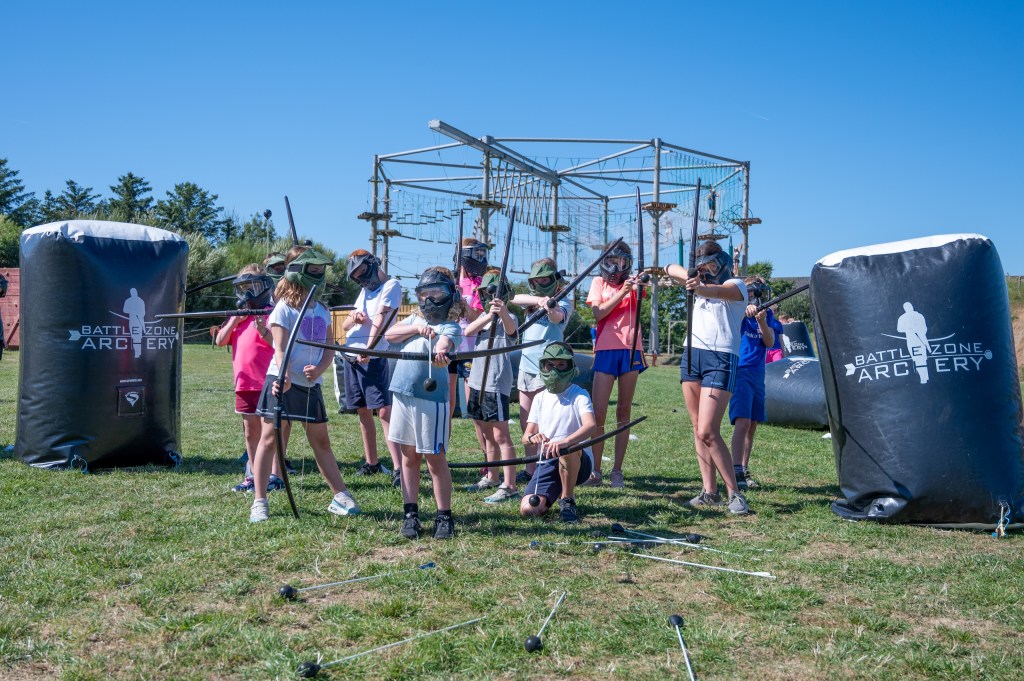 Children playing archery tag outdoors with safety gear.