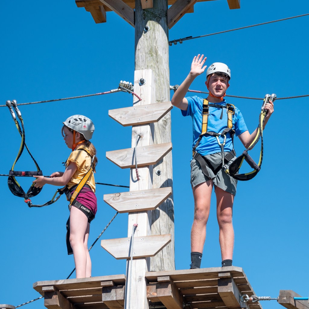 Kids in helmets on a ropes course, boy waving, girl adjusting harness.