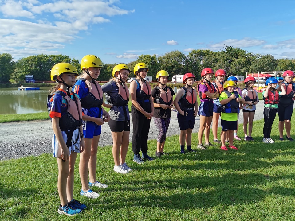 Group of people in helmets and life jackets standing on grass by a lake.