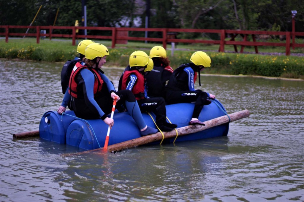 Group on blue raft in water, wearing yellow helmets and life jackets.