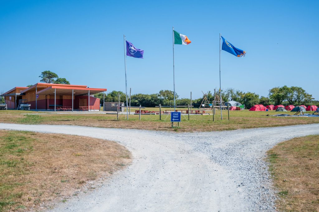 Three flags flying near a campsite with tents and a building under a clear blue sky.
