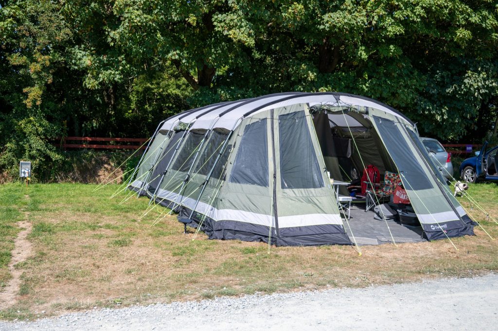 Large gray and green tent set up on grass near trees and a parked car.