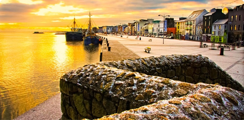 Sunset over a waterfront with boats, colorful buildings, and a stone path.