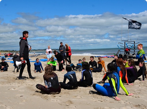 A group of people in wetsuits sitting on a beach, listening to a person speaking with a megaphone.