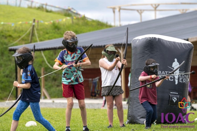 Children wearing masks play archery tag on a grassy field.