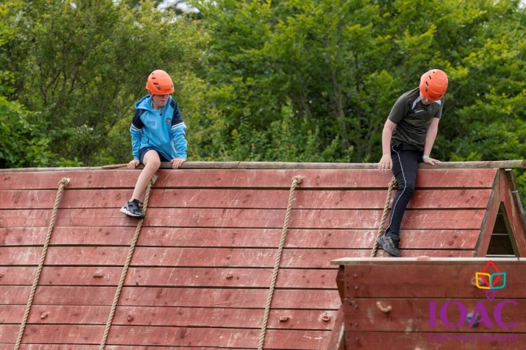 Two people in helmets climb a wooden obstacle wall outdoors.
