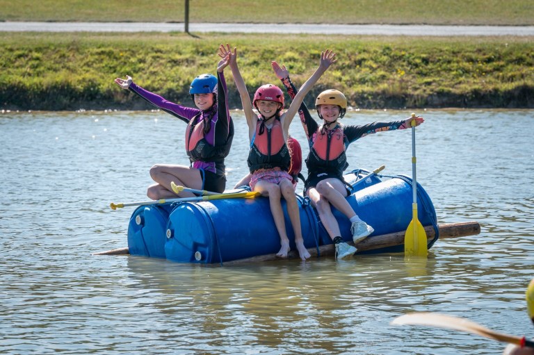 Three people in helmets and life vests sitting on a makeshift raft on a lake, raising their arms in excitement.