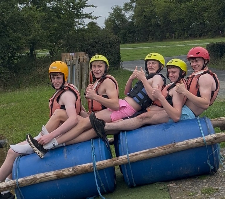 Five people in helmets sit on a makeshift raft, smiling and giving thumbs up.